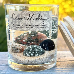May include: A clear glass jar candle with a bamboo lid. The candle is filled with a clear gel and has small rocks and sand at the bottom. The label on the jar reads "Lake Michigan" and "BERGAMOT | ROSEMARY MINT | WOODS".