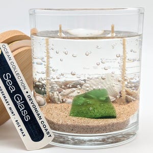 May include: A clear glass candle with a sea glass theme. The candle contains sand, seashells, and a green sea glass piece. The label reads "Sea Glass" and "Ocean Collection". The candle has two wicks and a wooden lid.