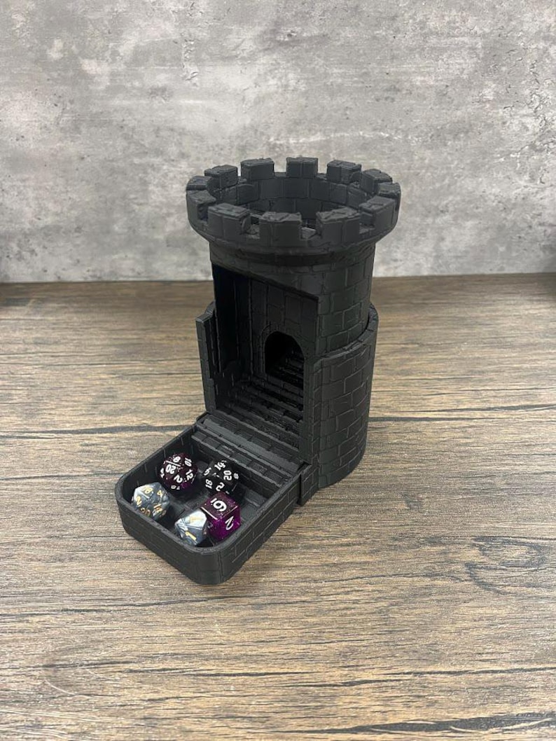 May include: A black 3D printed tower-shaped dice rolling tray with a hinged lid. The lid is open and reveals a set of dice inside.