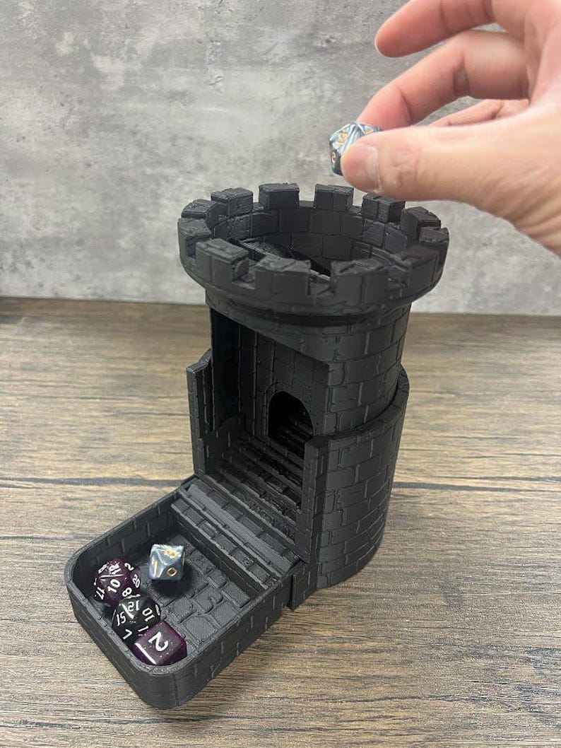 May include: A black 3D printed tower-shaped dice rolling tower with a removable tray. The tower has a brick pattern and a small opening at the top. The tray has a brick pattern and holds several dice.