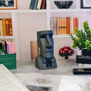 Peut inclure: Une statue Moai grise avec un nœud papillon, une boîte verte avec le texte "EVERYTHING IS FINE", une agrafeuse et une petite plante en pot. L'arrière-plan comprend des livres et des objets décoratifs.