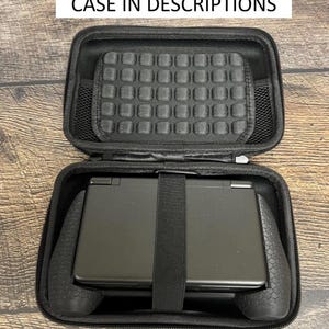 May include: A black zippered carrying case containing a black handheld gaming console with a textured grip. The interior lid features a padded grid section and a mesh pocket. Text at the top reads: LINK TO COMPATIBLE CASE IN DESCRIPTIONS.