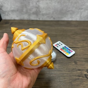 May include: A white and gold 3D printed orb with intricate designs. The orb is illuminated from within, creating a warm glow. The orb is being held in a hand.