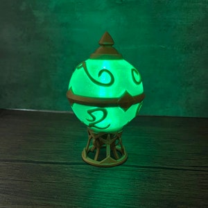May include: A green glowing orb with gold accents and intricate designs sits on a gold stand. The orb is illuminated from within, casting a soft green light.