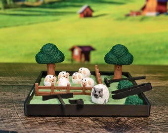 Puede incluir: Un jardín zen en miniatura con un marco negro, árboles verdes, una valla de madera y figuritas de ovejas blancas. Una figurita de lobo gris se encuentra en el centro del jardín.