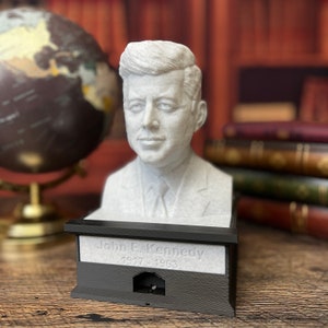 Könnte beinhalten: Eine weiße Büste von John F. Kennedy auf einem schwarzen Sockel mit der Inschrift "John F. Kennedy 1917 - 1963".
