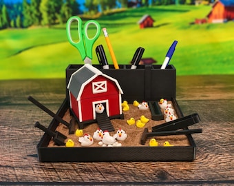 May include: A black desk organizer featuring a miniature red barn with a gray roof, a sand-filled base, and several small white and yellow chicken figurines. The organizer includes slots for stationery items like scissors, pens, and pencils.