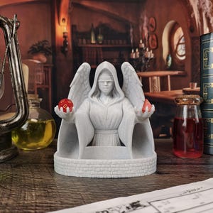 May include: A white statue of a robed figure with wings and a blindfold, holding a red die in each hand. The statue sits on a brick-like base. The background includes books, potions, and a lantern, suggesting a fantasy theme.