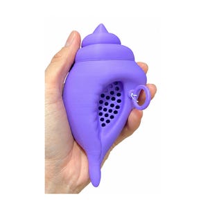 Puede incluir: Un infusor de té morado con forma de concha marina y acabado mate. El infusor presenta una superficie perforada en la parte frontal para preparar té en hebras e incluye un pequeño anillo circular en el lateral para facilitar su manipulación.