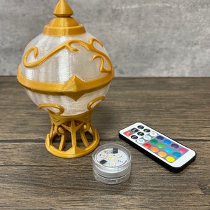 May include: A gold and white decorative lamp with a remote control for changing the color of the LED lights. The lamp is on a wooden surface.
