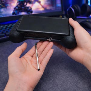 May include: A black handheld gaming console encased in a textured black grip, featuring a metallic silver telescopic stylus attached by a thin black cord. The device has a smooth, matte black exterior finish and is held in a horizontal orientation.