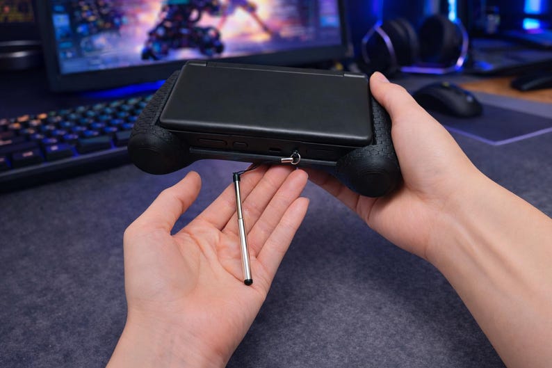 May include: A black handheld gaming console encased in a textured black grip, featuring a metallic silver telescopic stylus attached by a thin black cord. The device has a smooth, matte black exterior finish and is held in a horizontal orientation.