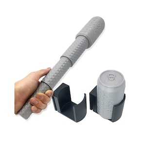 Puede incluir: Un porta latas de plástico gris con el texto "Woop Ass" impreso en él. El porta latas está diseñado para sostener una lata de aluminio de tamaño estándar. También se muestra en la imagen una varilla telescópica de plástico gris.