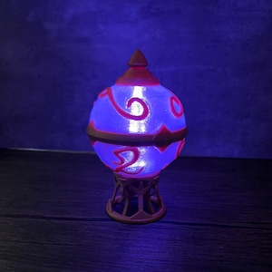 May include: A 3D printed lamp shaped like a glowing orb with intricate designs and a pink base. The orb is lit from within with a blue light.