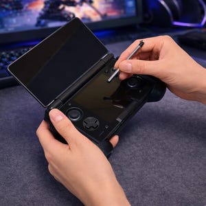 May include: A black handheld gaming console with a dual-screen design, featuring a top display and a bottom touchscreen interface. The device includes integrated directional pads, analog sticks, and buttons, accompanied by a thin metal stylus.