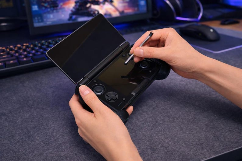 May include: A black handheld gaming console with a dual-screen design, featuring a top display and a bottom touchscreen interface. The device includes integrated directional pads, analog sticks, and buttons, accompanied by a thin metal stylus.