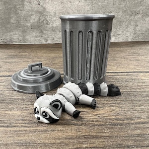 May include: A gray plastic trash can with a lid and a small gray raccoon figurine with black stripes lying next to it.
