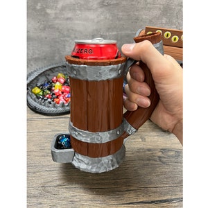 May include: A brown and silver beer stein-style mug with a handle, holding a red can of Zero. The mug has a built-in dice tray and is designed to look like a wooden barrel. A tray of colourful dice is in the background.