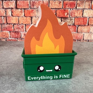 Könnte beinhalten: Ein grüner Müllcontainer mit einem Cartoon-Gesicht und dem Text "Everything is FINE" hält einen Holzflammen-Ausschnitt. Die Flamme hat orange und gelbe Schichten. Der Hintergrund ist eine Backsteinmauer.