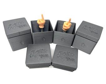 Könnte beinhalten: Sechs kleine schwarze Boxen mit dem Text "a gift for you" auf der Oberseite und einem federbelasteten Mittelfinger, der aus der Oberseite von zwei der Boxen herausspringt.