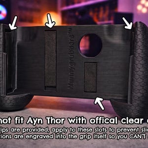 May include: A black handheld gaming controller grip with a textured honeycomb pattern on the sides. The center features the text "Mummygrubbers" and slots for foam strips. Text below states: "Cannot fit Ayn Thor with offical clear case. Foam strips are provided, apply to these slots to prevent sliding out. Instructions are engraved into the grip itself so you CAN'T MISS IT."