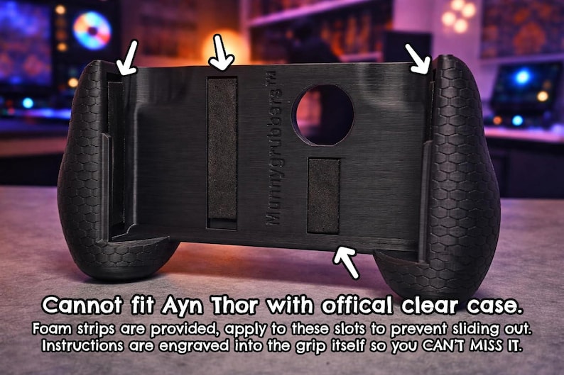 May include: A black handheld gaming controller grip with a textured honeycomb pattern on the sides. The center features the text "Mummygrubbers" and slots for foam strips. Text below states: "Cannot fit Ayn Thor with offical clear case. Foam strips are provided, apply to these slots to prevent sliding out. Instructions are engraved into the grip itself so you CAN'T MISS IT."