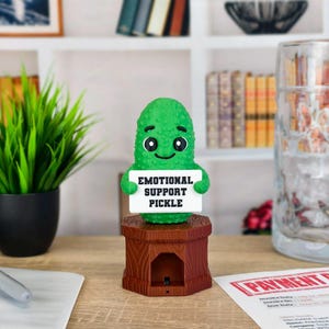 May include: A green pickle figurine with a smiling face holds a sign that reads "EMOTIONAL SUPPORT PICKLE." It sits on a brown wooden stand. A large glass beer mug and a small potted plant are in the background.