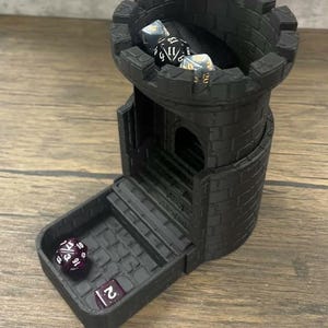 May include: Black plastic castle-shaped dice tower with a removable tray. The tower has a crenellated top and a small door. There are four dice inside the tower, and one die in the tray. The dice are various colors and have different numbers on them.
