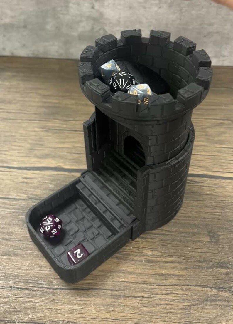 May include: Black plastic castle-shaped dice tower with a removable tray. The tower has a crenellated top and a small door. There are four dice inside the tower, and one die in the tray. The dice are various colors and have different numbers on them.