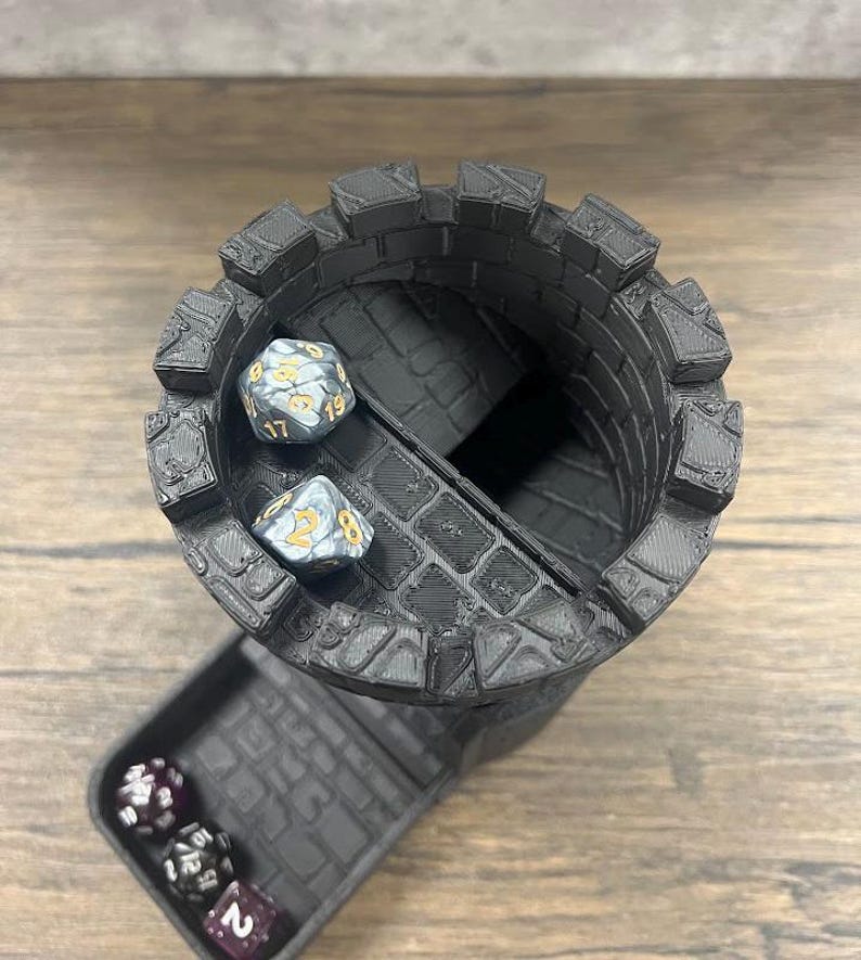 May include: Black 3D printed tower dice rolling tray with a removable top. The tower has a textured brick pattern. Two silver dice with gold numbers are in the tower. Three purple dice with white numbers are in the tray.