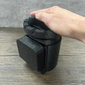 May include: Black castle-shaped dice tower with a square base. The tower is made of 3D printed plastic and has a brick pattern.