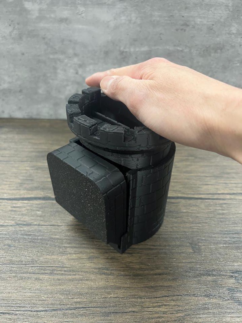 May include: Black castle-shaped dice tower with a square base. The tower is made of 3D printed plastic and has a brick pattern.