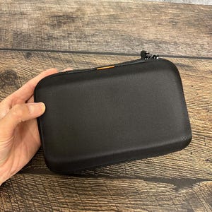 May include: A rectangular black hard-shell carrying case with a textured surface, rounded corners, and a zipper closure. A small orange accent detail is visible along the top edge near the zipper, which features two metal pull tabs.