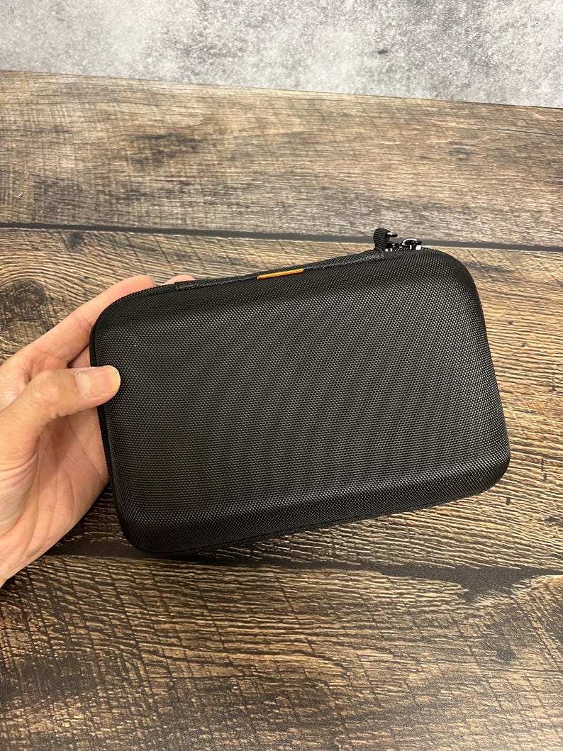 May include: A rectangular black hard-shell carrying case with a textured surface, rounded corners, and a zipper closure. A small orange accent detail is visible along the top edge near the zipper, which features two metal pull tabs.