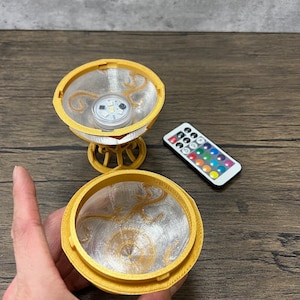 May include: A gold-colored decorative stand with a round top and a clear, textured dome. The stand has a small LED light inside the dome, which can be controlled by a remote control. The remote control has buttons for different colors and functions.