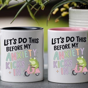 May include: Two white ceramic mugs with the text "Let's Do This Before My Anxiety Kicks In." One mug has a black handle and rim, the other has a pink handle and rim. Each mug features a green frog riding a pink scooter.