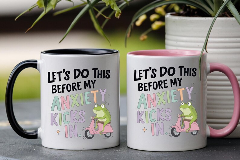 May include: Two white ceramic mugs with the text "Let's Do This Before My Anxiety Kicks In." One mug has a black handle and rim, the other has a pink handle and rim. Each mug features a green frog riding a pink scooter.
