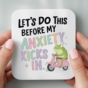 May include: Square white coaster with the text "LET'S DO THIS BEFORE MY ANXIETY KICKS IN." A cartoon green frog rides a pink scooter. The coaster is held by two hands with pink nail polish.