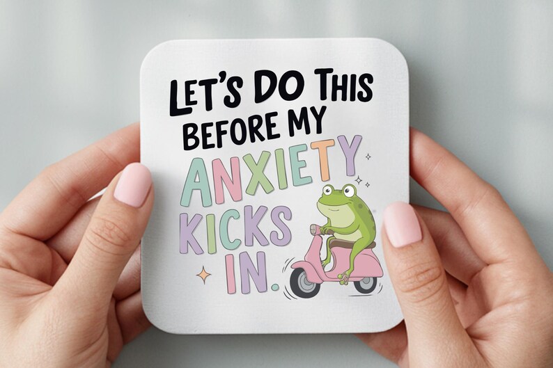 May include: Square white coaster with the text "LET'S DO THIS BEFORE MY ANXIETY KICKS IN." A cartoon green frog rides a pink scooter. The coaster is held by two hands with pink nail polish.