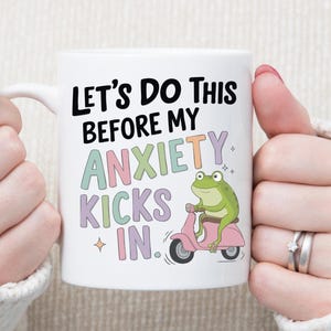 May include: White ceramic mug with the text "Let's Do This Before My Anxiety Kicks In." A green frog rides a pink scooter on the mug. The mug is held by a person wearing a white sweater.