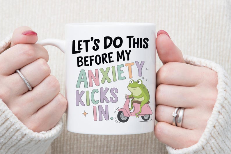 May include: White ceramic mug with the text "Let's Do This Before My Anxiety Kicks In." A green frog rides a pink scooter on the mug. The mug is held by a person wearing a white sweater.