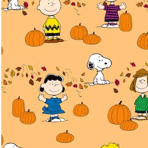 May include: A light orange fabric featuring a repeating pattern of Peanuts characters including Charlie Brown, Snoopy, Lucy, and Linus. The design includes various orange pumpkins and scattered autumn leaves in shades of red, brown, and yellow.