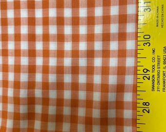 May include: Close-up of orange and white gingham fabric. The fabric is a classic checkered pattern, with evenly spaced squares of orange and white. A yellow ruler is visible along the right edge, with measurements in inches.