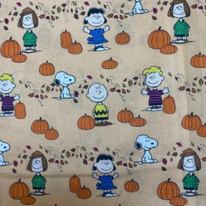 Może przedstawiać: Beżowy materiał z powtarzającym się wzorem postaci z Peanuts i dyni. Postacie to Charlie Brown, Snoopy, Lucy i Linus. Dynie są pomarańczowe i mają łodygę.