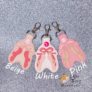 May include: Three keychains with embroidered ballet shoe designs. The keychains are beige, white, and pink. The keychains are labeled "Beige", "White", and "Pink".