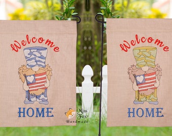 May include: Two burlap welcome home flags with a gnome holding an American flag. The gnome on the left is wearing a blue and white camouflage outfit and the gnome on the right is wearing a green and yellow camouflage outfit. Both flags have the text "Welcome Home" and a fox logo with the text "Miss Foxy Handmade".