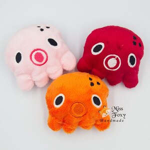 May include: Three plush octopus toys in pink, red, and orange. Each has large black eyes and a central circular design. The toys are handmade and have a soft, fuzzy texture. The octopus toys are a cute and whimsical design.