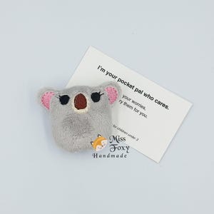 May include: A grey plush koala bear with pink ears and a brown nose, next to a card that reads "I'm your pocket pal who cares." The card also has the text "Miss Foxy Handmade."