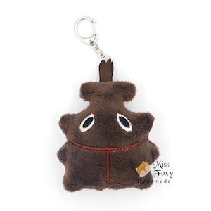 Puede incluir: Un llavero de peluche marrón con forma de animal estilizado, con ojos blancos y negros y un contorno cosido en rojo. El llavero tiene un cierre de metal plateado y un lazo de cinta marrón.