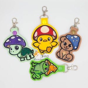 May include: Four embroidered keychains featuring cartoon animals: a turtle with a purple mushroom cap, a yellow chick with a red and white mushroom, a brown dog with a blue mushroom, and a green frog with an orange mushroom. Each keychain has a metal clasp.